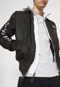 Giacca bomber nera con esterno lucido, polsini a costine e cerniere. Indossata sopra un top grigio chiaro, con un patch del logo e dettagli bianchi.