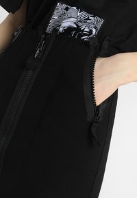 Svart plagg med flera dragkedjor, en texturerad design och en kontrasterande ficka med ett monokromt abstrakt mönster.