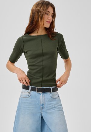 Noor naine, kes kannab oliivirohelist lühikeste varrukatega tihedalt istuvat topi ja helerohelisi laia säärega kõrge vöökohaga pükse, kohandab oma musta vööd.