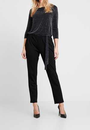 Kobieta w czarnych spodniach z wysokim stanem, zwężanych u dołu, czarnych szpilkach z ostrym noskiem oraz połyskującej bluzce z długim rękawem i ozdobnym wiązaniem po boku.