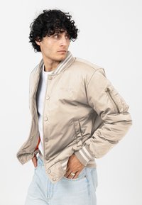 Beżowa kurtka bomberka o satynowym wykończeniu, z prążkowanymi mankietami i kołnierzem; posiada kieszeń boczną z zamkiem błyskawicznym oraz detal z logo na klatce piersiowej.