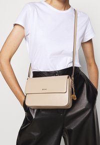 Béžová kožená kabelka přes rameno se zapínáním na klopu, zlatým logem a řetízkovým detailem, nošená přes bílé tričko a černé kalhoty s vysokým pasem.