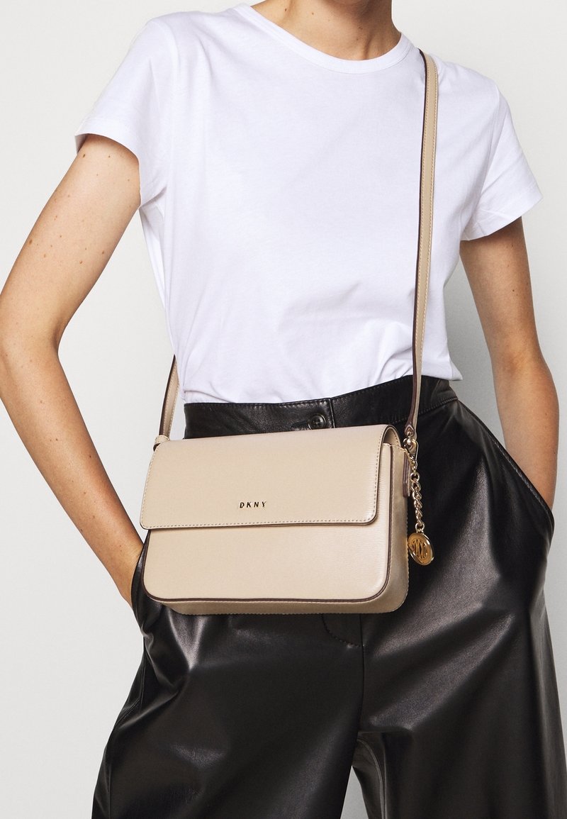 Béžová kožená kabelka přes rameno se zapínáním na klopu, zlatým logem a řetízkovým detailem, nošená přes bílé tričko a černé kalhoty s vysokým pasem.