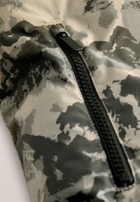 Fekete cipzár szürke és fekete álcázott anyagon, részben kinyitva, látható anyagtextúrával és varrással.