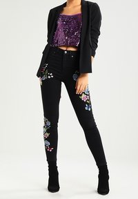 Jean noir avec broderie florale colorée sur les côtés, associé à un crop top violet scintillant et un blazer noir cintré. Bottines noires.