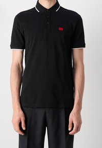 Svart polo skjorta med ribbad krage, treknappat knäppning, vita detaljer på ärmarna och en liten röd logotyp på bröstet.