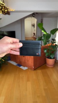 Porta carte di credito nero con texture a coste, molteplici scomparti per carte e un piccolo logo. Tenuto in mano, con un pavimento in legno e piante da interno sullo sfondo.