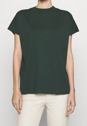 Osoba ubrana w jednokolorowy ciemnozielony t-shirt z krótkim rękawem i jasnobeżowe spodnie, stojąca na jednolitym tle.