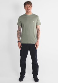 T-shirt en coton vert olive à manches courtes avec un petit logo sur la poitrine, associé à un pantalon en velours côtelé noir et des chaussures noires.