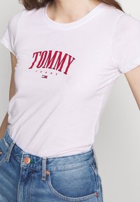 White short-sleeve t-shirt with a red "TOMMY JEANS" logo and small blue and red detail at the hem, paired with high-waisted blue denim jeans.