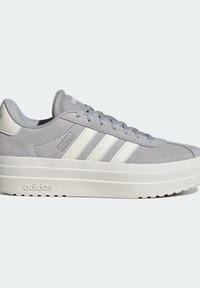 Grå mockasinsneaker med vit gummisula, med tre vita ränder på sidan, rund tå och snörning.