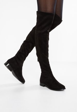 Schwarze Overknee-Flachstiefel aus Wildleder, getragen mit transparenten schwarzen Strumpfhosen, mit seitlichem Reißverschluss und niedrigem Blockabsatz vor weißem Hintergrund.