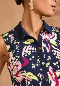 Majica brez rokavov v temno modri barvi s pisanimi cvetličnimi motivi v roza, zeleni in beli barvi. Ima ovratnik in rjave gumbe kot detajl.