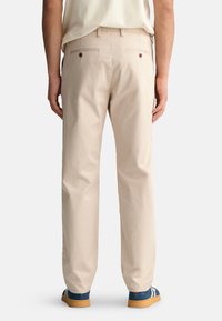 Beige Baumwollhosen mit geradem Schnitt, ausgestattet mit aufgesetzten Gesäßtaschen, die mit braunen Knöpfen gesichert sind. Dazu werden blaue und weiße Sneakers getragen.