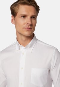 Bela srajca z gumbom in ostrim ovratnikom, žepom na levi strani prsnega dela in bisernimi gumbi. Gladko blago z ostrim videzom.