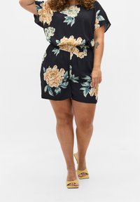 Frau trägt einen marineblauen floral bedruckten Kurzarm-Overall und gelbe Open-Toe-High-Heels, steht mit einem erhobenen Arm vor einem einfarbigen Hintergrund.