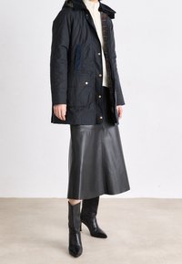 Osoba oblečená v černém kapucínovém kabátu, bílém pleteném svetru, černé kožené midi sukni a černých botách na podpatku stojí na béžové podlaze.