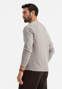 Uomo con barba e capelli corti ricci, che indossa una camicia a maniche lunghe grigia chiara e pantaloni scuri, che guarda di lato su uno sfondo bianco neutro.