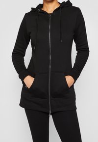 Sweat à capuche noir avec fermeture éclair, capuche, manches longues et poches avant. Confectionné dans un tissu lisse et extensible avec des poignets et un ourlet côtelés.