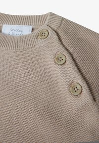 Beige gestrickter Pullover mit ribbelndem Ausschnitt, zwei runden Knöpfen an der Schulter und einer weichen Textur. Etikett lautet "Stellou & friends."