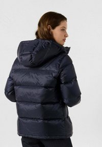 Mujer vestida con una chaqueta acolchada con capucha negra, de perfil contra un fondo claro y liso.