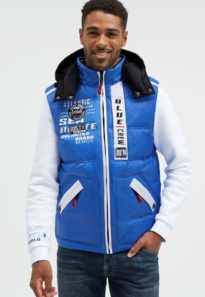 Blau gesteppte Weste mit schwarzem Kragen, weißen Akzenten und mehreren Reißverschlüssen. Mit aufgedruckten Logos und Mustern auf der Vorderseite. Getragen über einem weißen Langarmshirt.