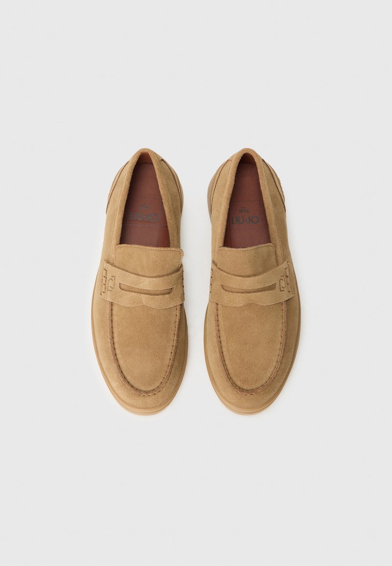 Mocasines de ante marrón, sin cordones, con punta redondeada, que cuentan con una tira decorativa y detalles de costura en la parte superior. Suela de goma en contraste.