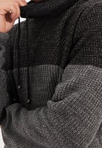 Gebreide pullover met een hoge kraag in grijs en zwart, voorzien van een gestructureerd patroon en zwarte koordjes voor een verstelbare pasvorm.
