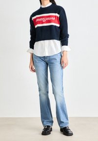 Persona vistiendo un suéter corto azul marino con el logo "Tommy Jeans", una camisa blanca de botones, jeans de color azul claro y mocasines negros, de pie sobre un fondo liso.