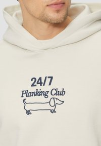 Svetlobéžová mikina s kapucňou a predným dizajnom, na ktorom je nápis "24/7 Planking Club" tmavomodrou farbou a jednoduchý obrys jazvečíka.