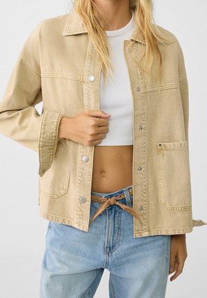 Kvinde iført beige knapjakke over hvid cropped top og lyseblå jeans med tan bælte bundet foran.