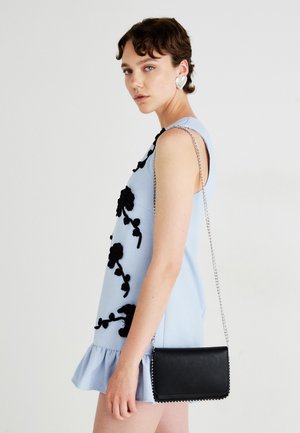 Hellblaues ärmelloses Kleid mit schwarzer floralem Stickerei, rüschigem Saum und einer kleinen schwarzen Umhängetasche mit Kettenriemen.