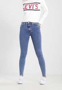 Jeans skinny de mezclilla azul con cintura alta, que presentan un diseño clásico de cinco bolsillos y sutilezas en la costura. Combinados con una blusa blanca de manga larga.