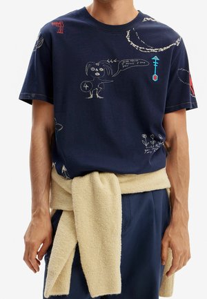 Maglietta blu navy con scarabocchi bianchi, rossi e blu, inclusa una persona che tiene uno scudo e un fumetto. Maglione beige legato in vita.