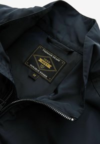 Donkerblauwe jas binnenkant met zwart label. Gouden letters met de tekst "PREMIUM GRADE 1982 NEXT DURABLE GOODS," maatlabel "M." Ritsopening.