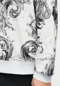 Κοντινό πλάνο λευκού φούτερ με μαύρα και γκρι στροβιλιζόμενα σχέδια κυμάτων και πιτσιλιές, φορεμένο με μαύρο παντελόνι, που δείχνει λεπτομέρειες στον καρπό και το τελείωμα.