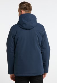 Tamsiai mėlyna vandeniui atspari striukė su kapišonu, pasižyminti lygiu paviršiumi, ilgaamžėmis rankovėmis ir tiesiu apvadu. Nugarinė pusė rodo siuvimo detales.