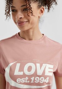 Lichtroze katoenen t-shirt met witte grafische tekst "LOVE est. 1994" en logo. Korte mouwen, ronde halslijn, zachte textuur.
