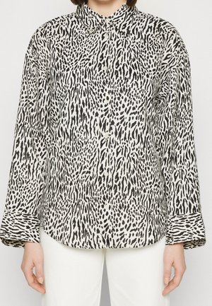 Femme portant une chemise à manches longues boutonnée avec un imprimé animal abstrait noir et blanc, associée à un pantalon blanc.