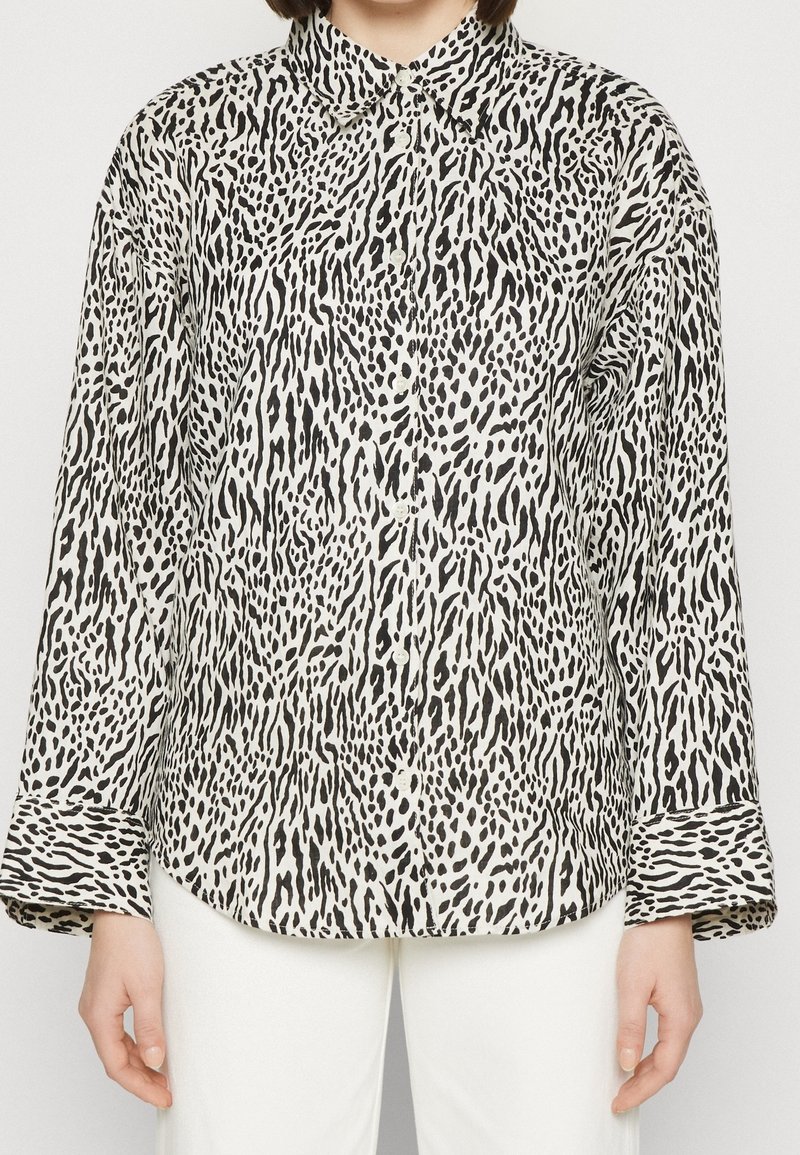 Femme portant une chemise à manches longues boutonnée avec un imprimé animal abstrait noir et blanc, associée à un pantalon blanc.