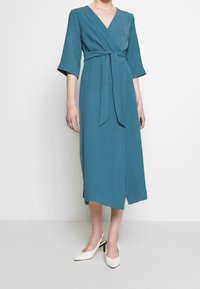 Robe midi teal à manches 3/4 et ceinture à nouer, portée avec des escarpins slingback blancs à bout pointu, se tenant devant un fond uni.