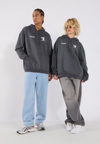 To personer står skuldre ved skuldre i hettegensere. Den grå hettegenseren har en frontlomme og trykte logoer. De har på seg forskjellige stiler av baggy jeans og sneakers.