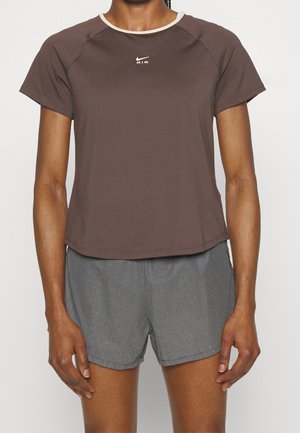 Frau in einem braunen Nike Air T-Shirt mit kurzen Ärmeln und grauen Sportshorts, die vor einem schlichten weißen Hintergrund steht.