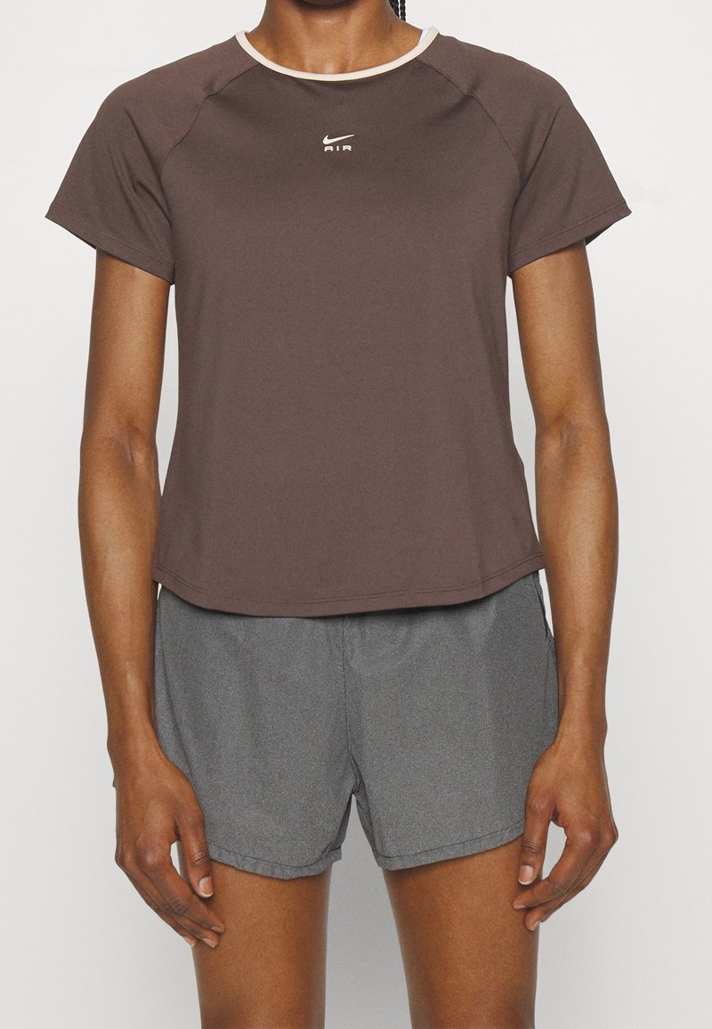 Person som bär en brun Nike Air t-shirt med kort ärm och grå sportshorts, visad från halsen till halva låret mot en enkel bakgrund.