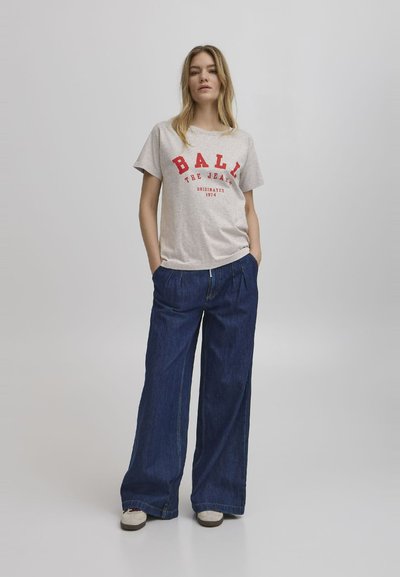 Gaišpelēka kokvilnas T-kreklu ar sarkanu uzrakstu "BALL THE JEANS 1974", apvienots ar platajiem tumši zilajiem džinsiem un baltajām kedām.