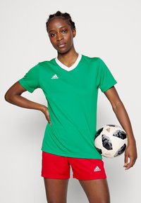 Zelené športové tričko s bielym golierom, v kombinácii s červenými šortkami. Hráč drží čierno-biely štýlový futbalový loptu. Viditeľné logo Adidas.