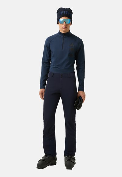 Haut de sport navy avec un zip demi, logotype noir, associé à un pantalon navy avec deux boutons. Le mannequin porte des gants noirs et des lunettes de soleil miroir.
