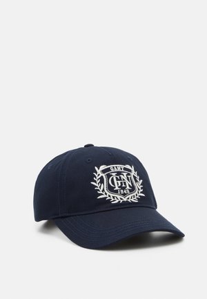 Marineblå baseballkasket med buet skygge, med hvidt broderet våbenskjold og teksten "GANT 1949" på frontpanelet.
