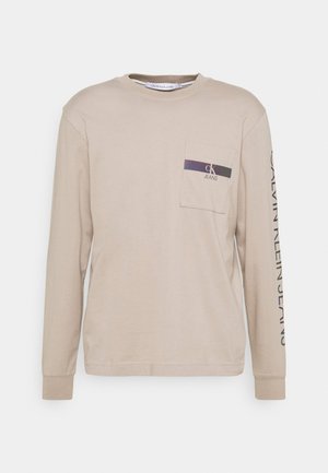 Beige Langarm-T-Shirt aus weichem Baumwollstoff mit einer Tasche, die einen lila und schwarzen Logodruck sowie einen Markentext entlang eines Ärmels aufweist.
