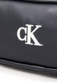 Calvin Klein sötétkék bőr táska texturált felülettel és fehér hímzett 'ck' logóval az elején. Négyzet alakú, minimális hardverrel.